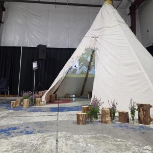 A 30-foot tall tipi decorated with fireweed and wood logs around. Inside, two projectors mapped along the tipi's canvas displaying vibrant, immersive visuals.